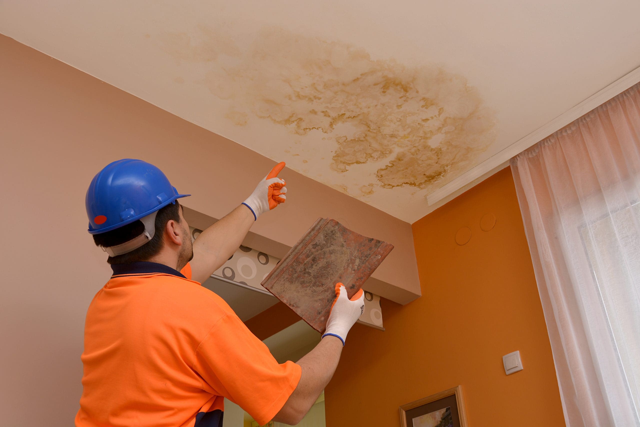 A worker wearing an orange shirt and blue helmet inspects a large, dark water stain on a white ceiling. He points toward it while holding a trowel. The room has light pink walls and a window with sheer curtains.