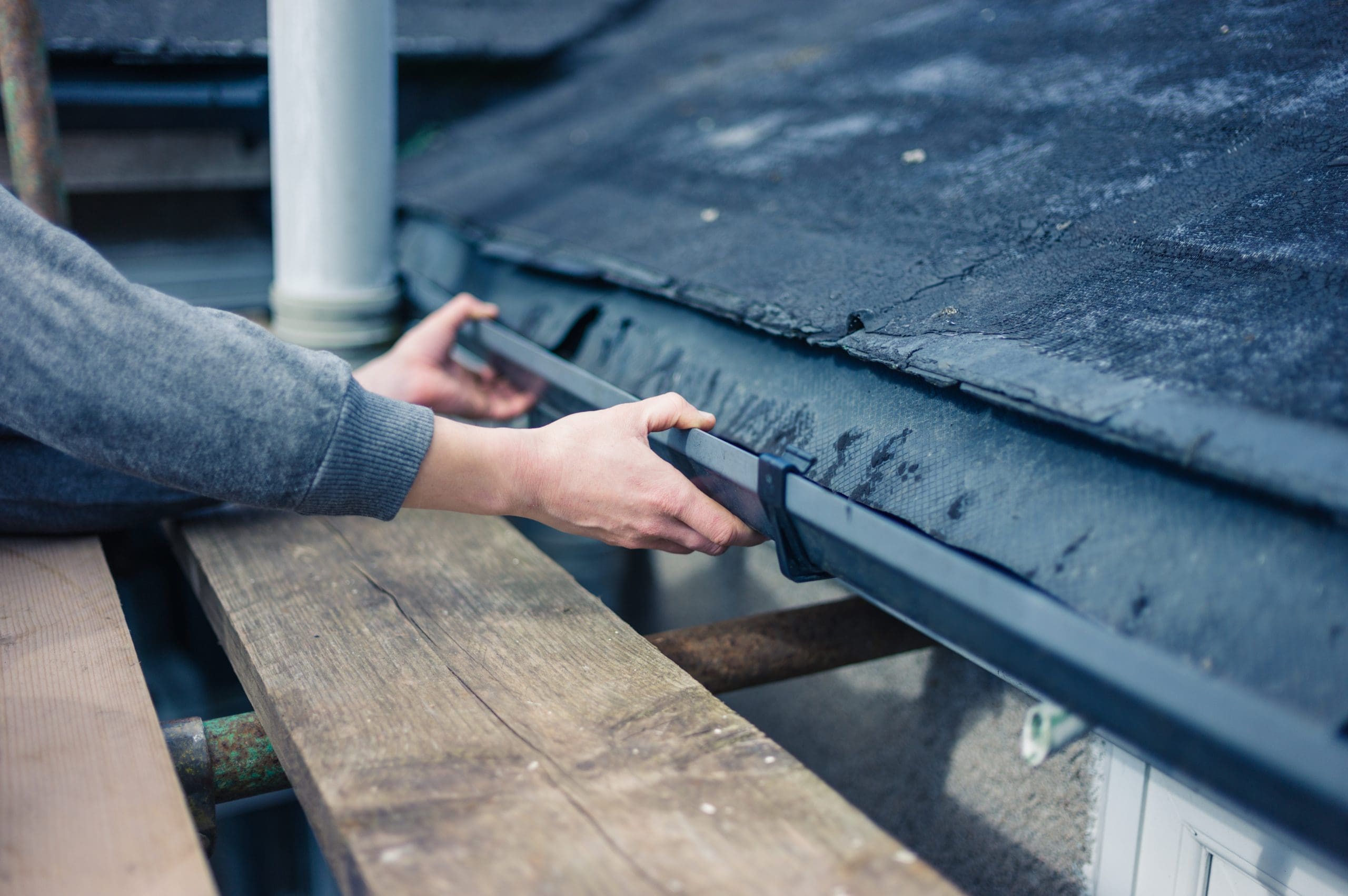 A person wearing a gray sweater is installing or adjusting a black gutter on the edge of a roof. The individual is standing on wooden scaffolding, and the roof appears to have weathered shingles.