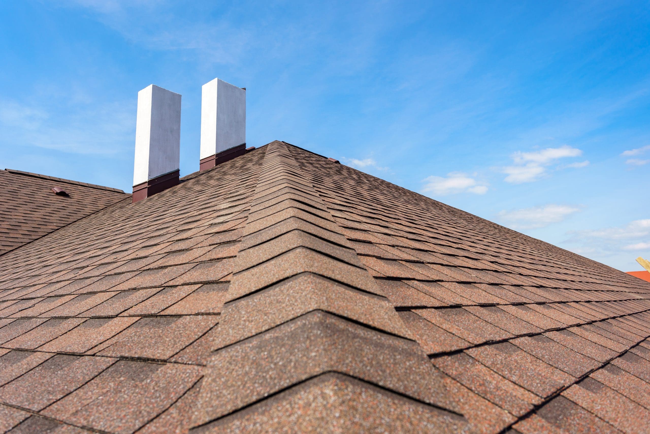 A close-up view of a rooftop with brown asphalt shingles, featuring two white chimneys under a clear blue sky. The roofline stretches diagonally from the bottom left to the top right of the image.