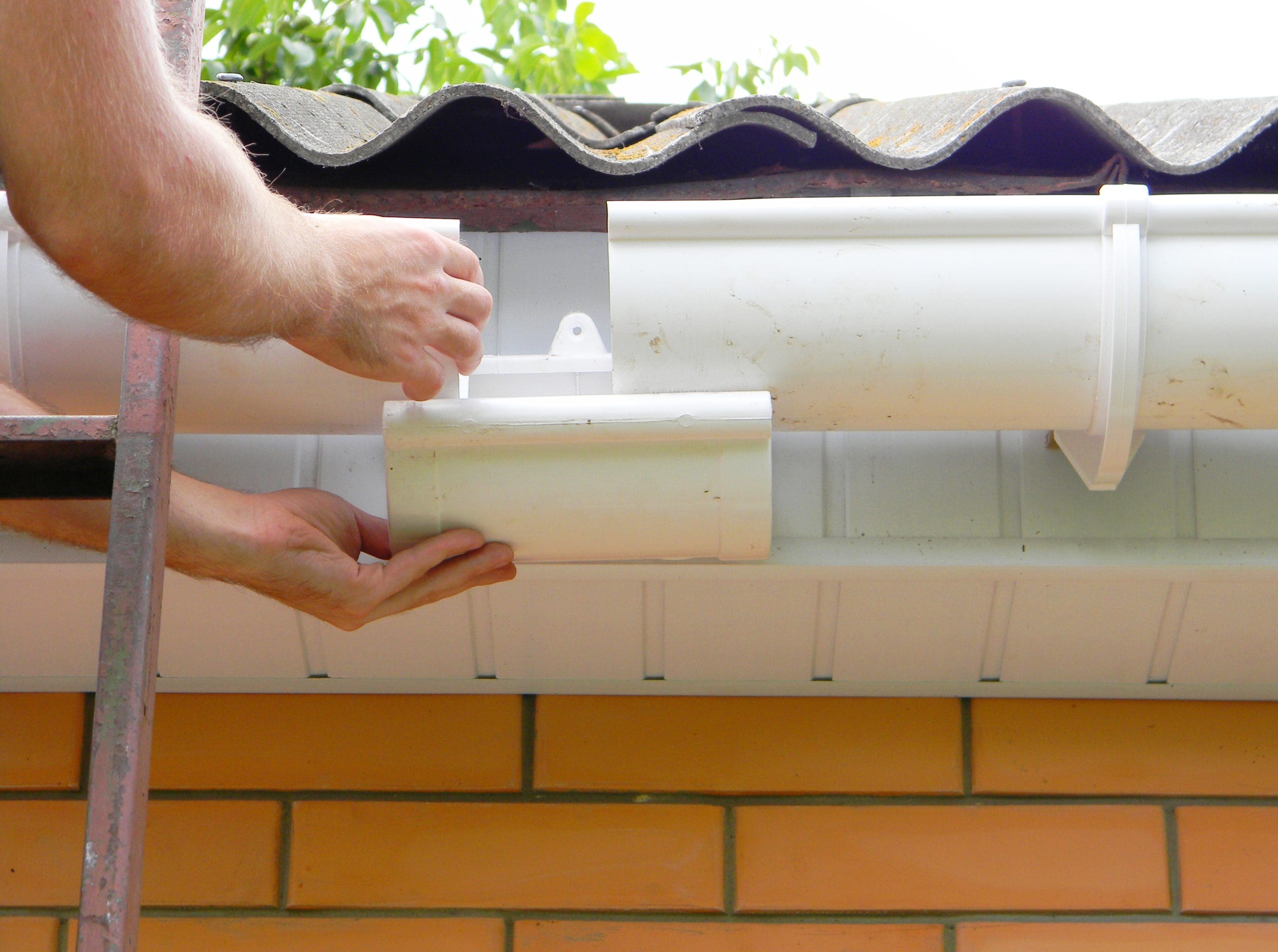 A person is installing or repairing a white plastic gutter system on a building with brick walls. One hand holds part of the gutter while the other hand secures a section. The roof has corrugated tiles, and a ladder is visible.