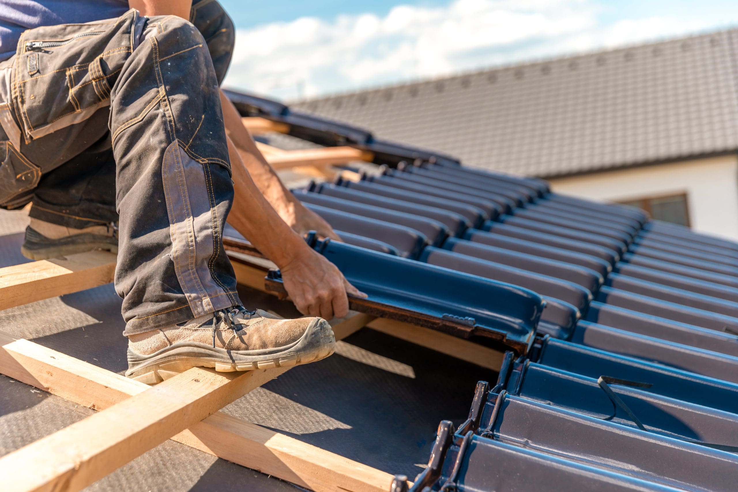 A person wearing work boots and jeans is installing dark roofing tiles on a house roof. The sky is clear and the background shows part of a completed tiled roof.