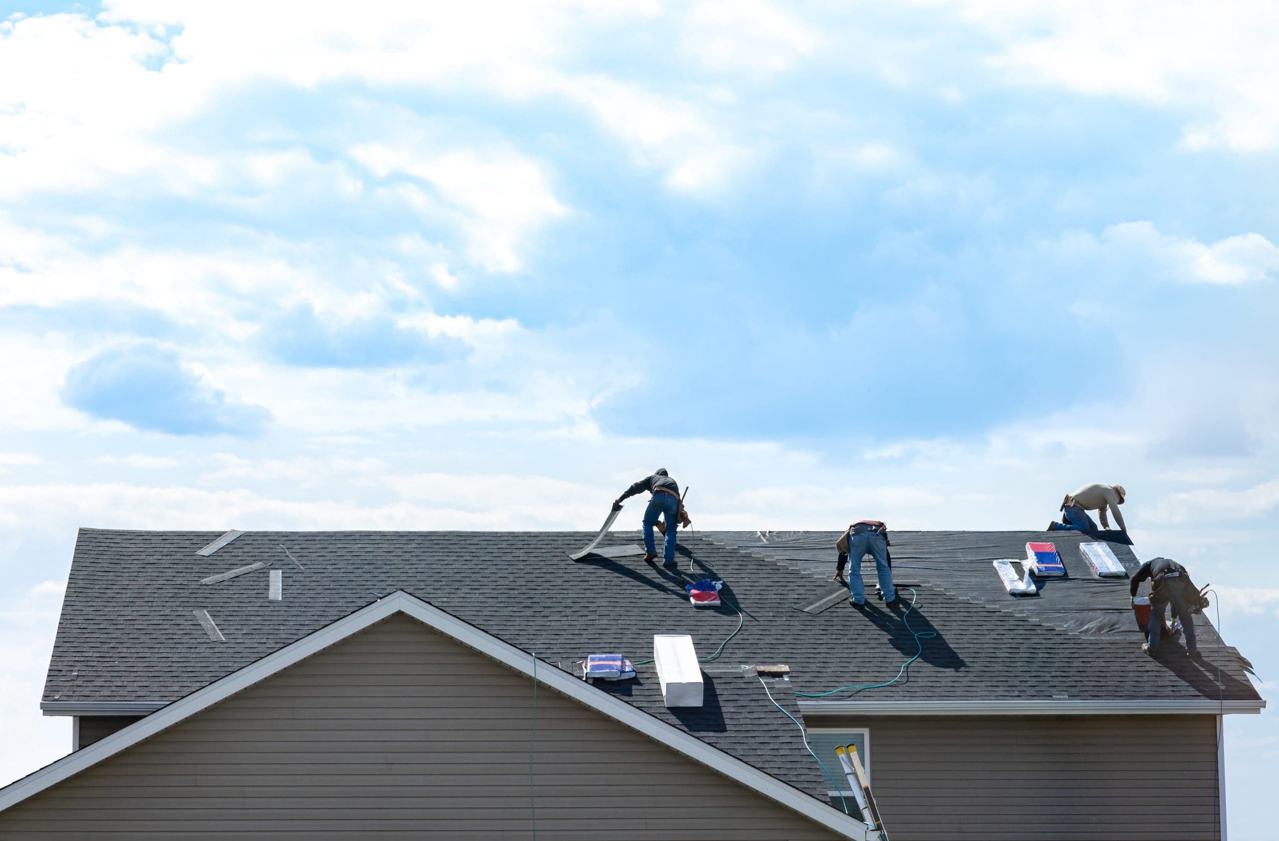 Workers on a roof installing shingles on a residential building under a partly cloudy sky. Various tools and materials are scattered around as they work. The roof is partially completed, with some sections still uncovered.