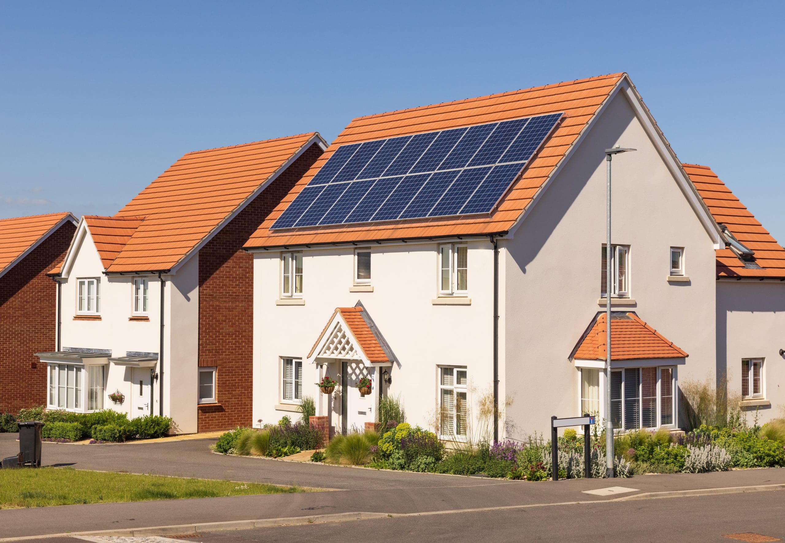 A row of modern suburban houses with red-tiled roofs on a sunny day. The house in the foreground has solar panels installed on the roof. Well-maintained lawns and flower beds line the street, and a streetlamp is visible nearby.