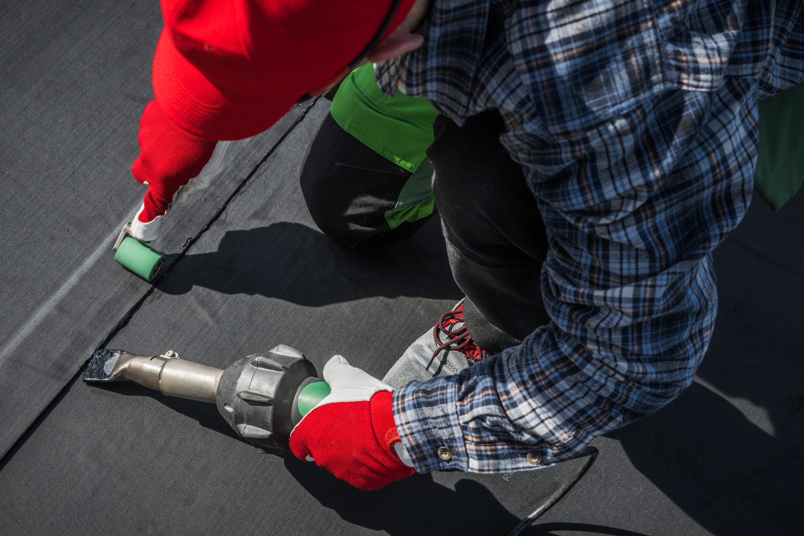 A person wearing a red hat and gloves, a plaid shirt, and black pants uses a heat-sealing tool to weld the seam of a black membrane on a flat roof. They are kneeling and working carefully on the surface.