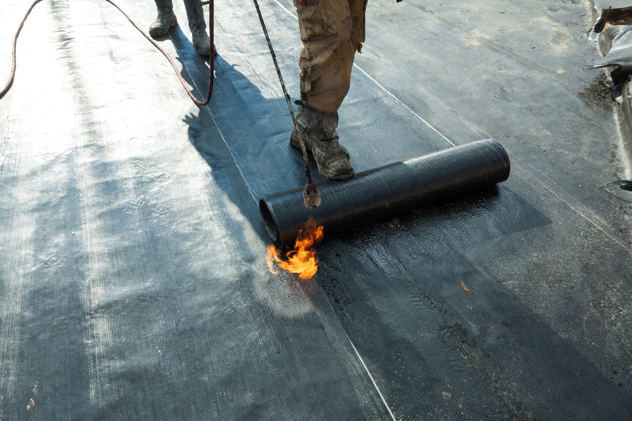 A worker uses a torch to unroll and melt a roofing membrane onto a flat roof surface. The membrane is being heated to adhere it securely in place, with flames visible from the torch. The worker is wearing protective boots.