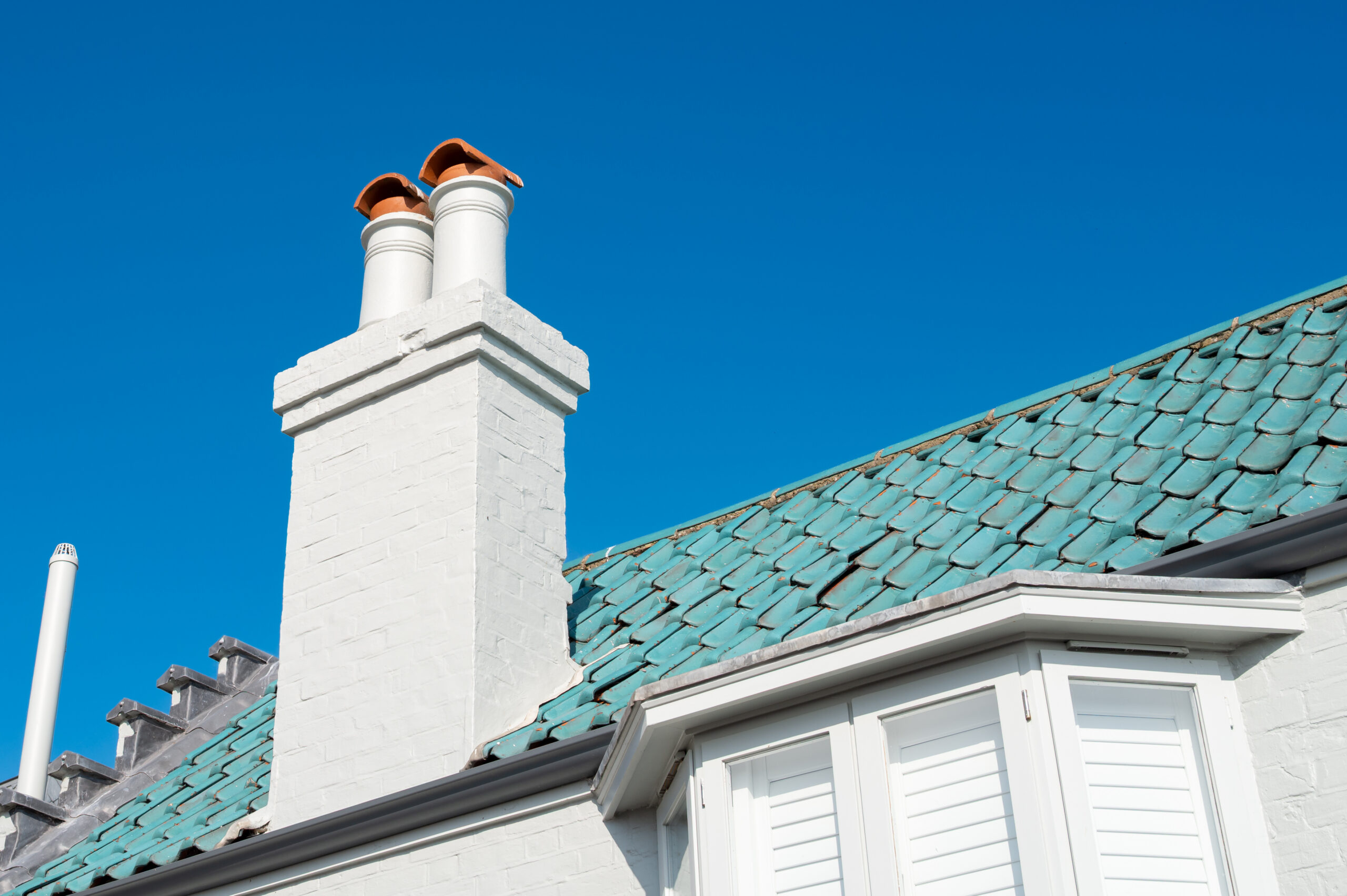 A white brick chimney with two clay-topped flues rises above a house with a green tiled roof and closed white shutters, set against a clear blue sky.