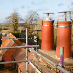 Two red brick chimney pots with metal cages on top of a rooftop, surrounded by scaffolding. In the background are trees, houses, and a road on a sunny day.